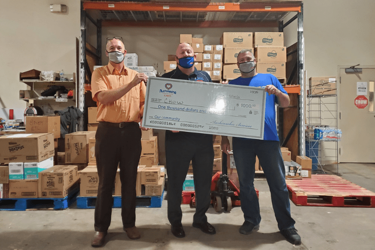 Three people stand proudly in a storage area, holding a large $1000 check. Shelves filled with cardboard boxes are visible behind them, showcasing their dedication to organized growth. This moment reflects more than just success; it's about us coming together to achieve shared goals.