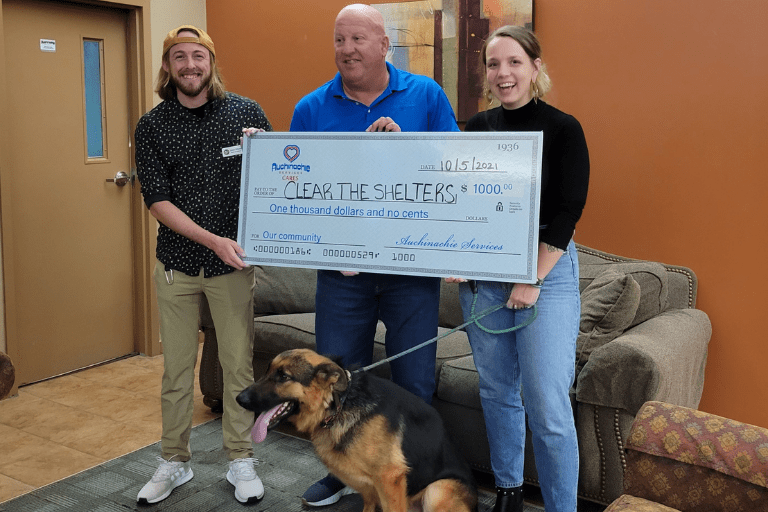 Three smiling individuals hold a large novelty check for $1000 addressed to "Clear the Shelters," with a proud German Shepherd standing in front of them, embodying our mission. Learn more about us and our commitment to making a difference in the lives of animals.