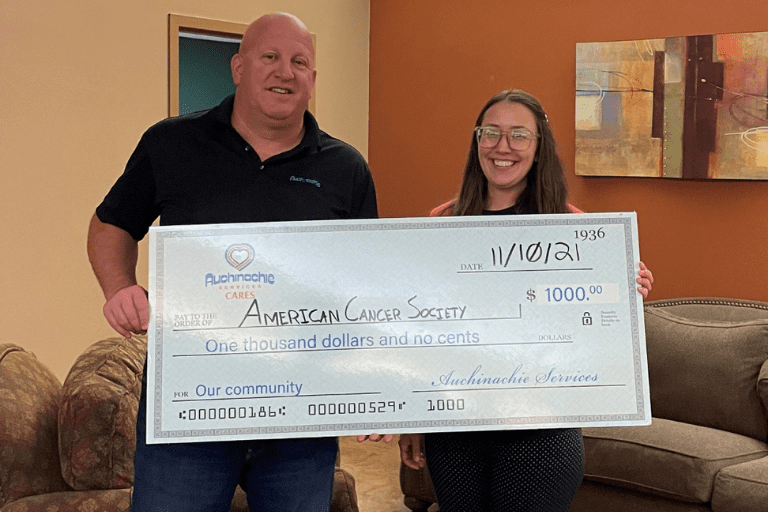In a warm room adorned with artwork, two individuals proudly hold a large $1000 check made out to the American Cancer Society, dated 11/10/21. This moment speaks volumes about us and our commitment to supporting impactful causes.