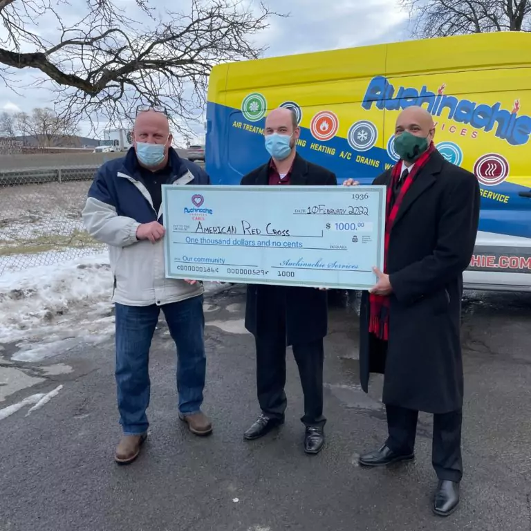 Three people stand outdoors holding a large check for the American Red Cross, amounting to $1000. A service van with the "About Us" section prominently displayed is parked in the background.