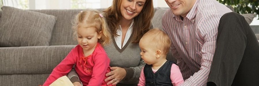 A family of four sits on a living room floor, engaging with a book. Parents are smiling, and the children, one toddler and one young child, are looking at the book.