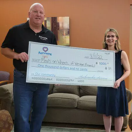 Two people stand indoors holding a large check for $1000, dated 9/1/22, from Auchinachie Services to Meals on Wheels of Western Broome.