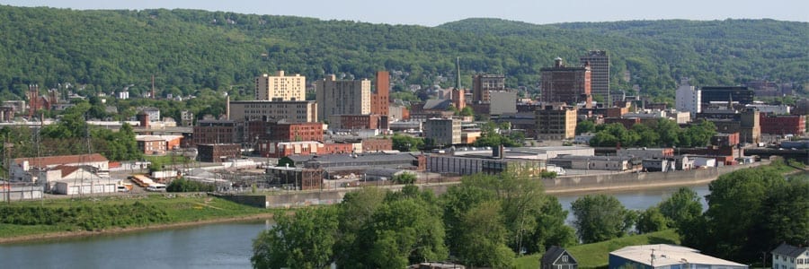 Panoramic view of a cityscape with a river in the foreground, surrounded by trees and hills in the background. Buildings of various heights are visible under a clear sky.