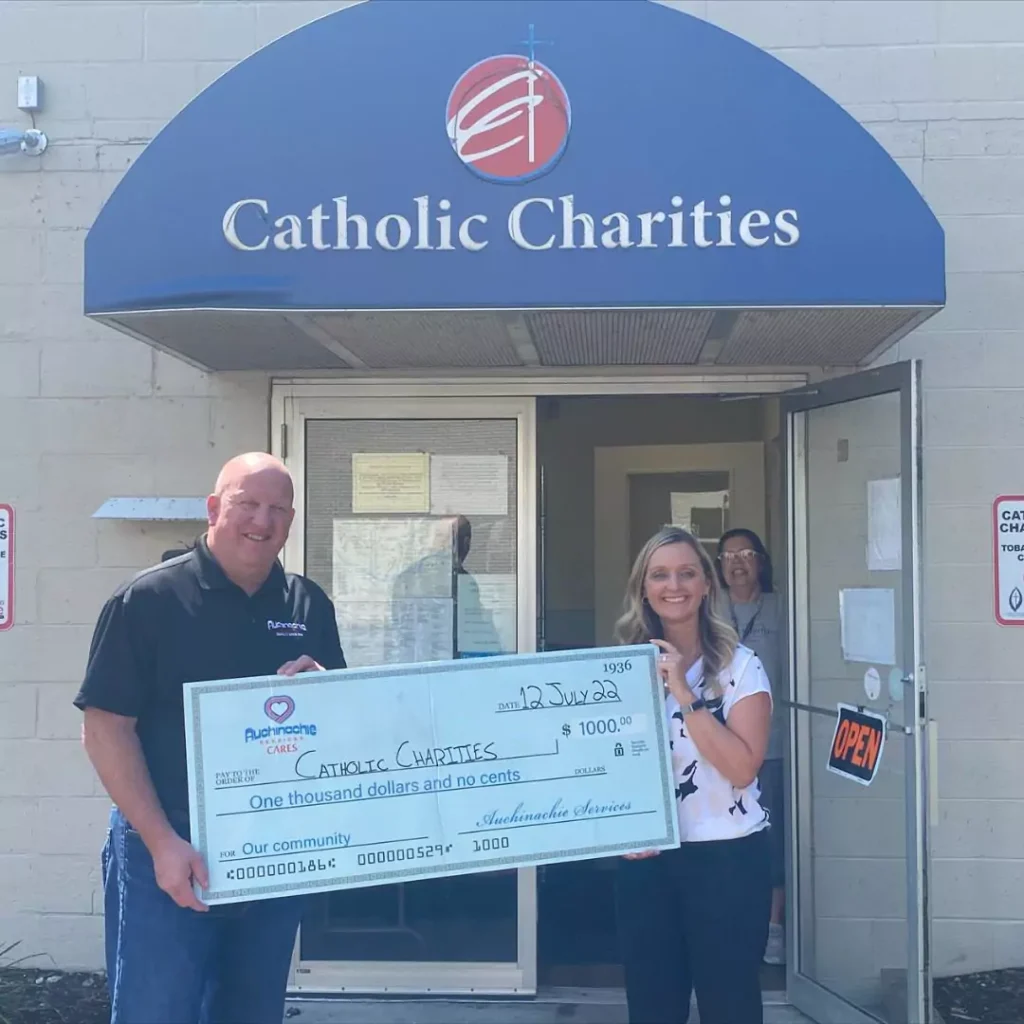 Two people stand in front of Catholic Charities holding a large check for $1,000, highlighting their commitment to giving back. An additional person is visible in the doorway, embodying the spirit of the About Us mission to foster community support and involvement.