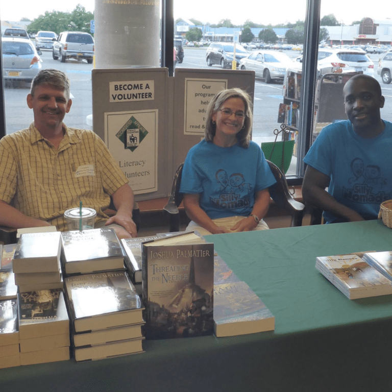 Three people seated at a table with stacks of books and a sign reading "Become a Volunteer." A window shows a parking lot outside, inviting passersby to learn more about us and join the cause.