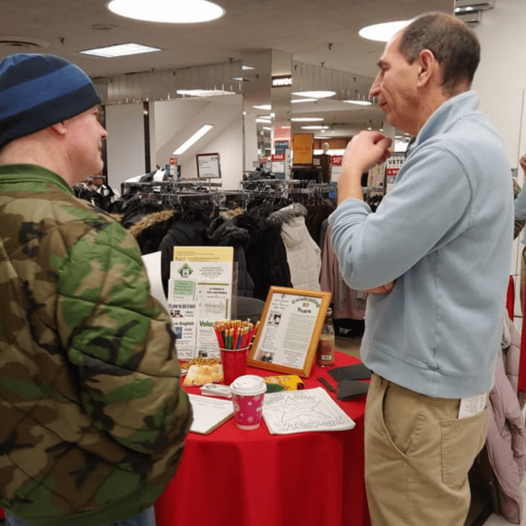 Two men chat at a table with brochures and colorful papers in a store. One sports a camo jacket and beanie, the other a light blue sweater. Coats are displayed in the background, setting the scene like an inviting "About Us" page come to life.