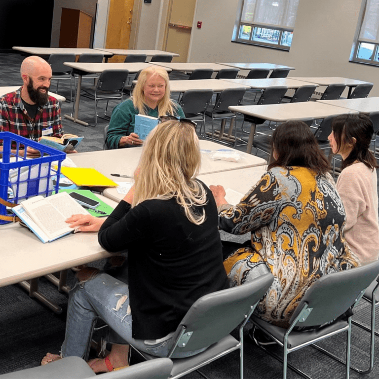 Five people sit around tables in a cozy classroom setting, engaging in a book discussion that feels as intimate as an "about us" page unfolding in real-time.