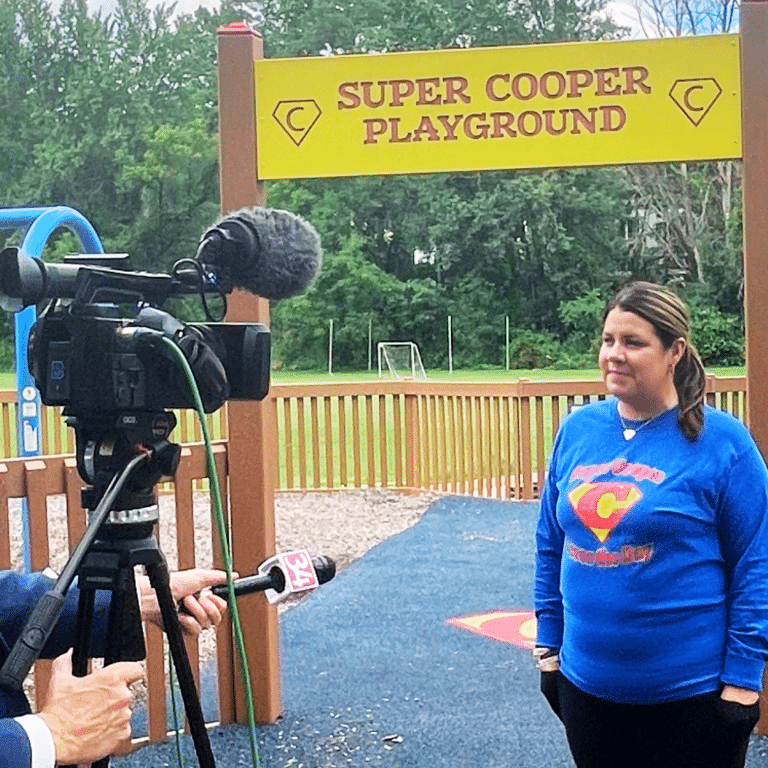 A woman in a blue shirt is being interviewed by a TV crew at the "Super Cooper Playground." A camera and microphone are focused on her.