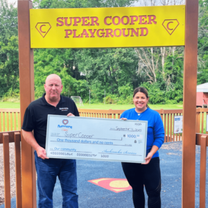 Two people hold a large check for one thousand dollars in front of a sign that reads "Super Cooper Playground." Trees and playground equipment are visible in the background.