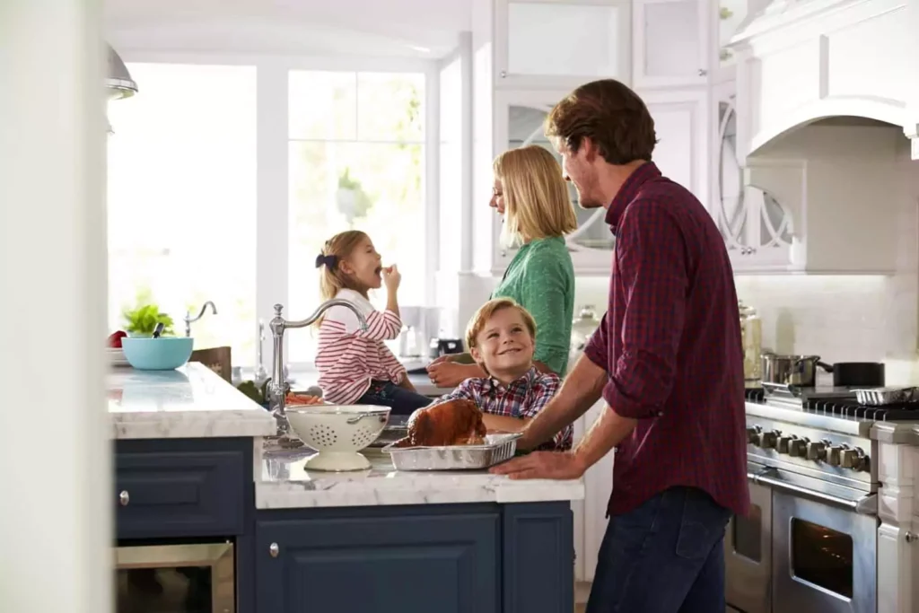 A family of four gathers in a kitchen. Two adults stand by the counter, one with a dish, while a child enjoys a snack and another looks at an adult. The kitchen is bright and modern.