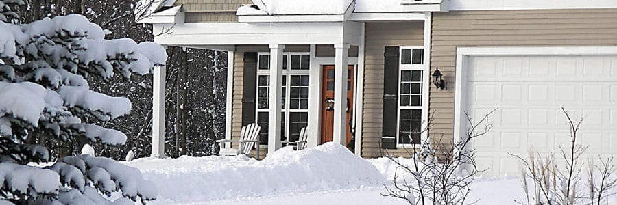 A snow-covered house with a porch, two chairs, and a garage. Snow blankets the roof, trees, and ground around the home.
