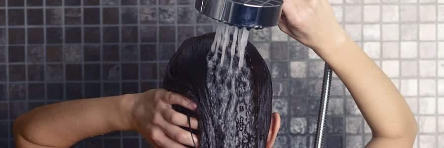 Person showering, water flowing from showerhead onto their head, with hands adjusting hair. Background shows dark tiled wall.