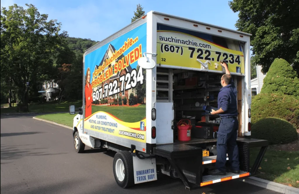 A person stands at the back of a plumbing service truck with open doors, accessing equipment. The truck displays contact information and advertises solutions for plumbing, heating, and air issues.