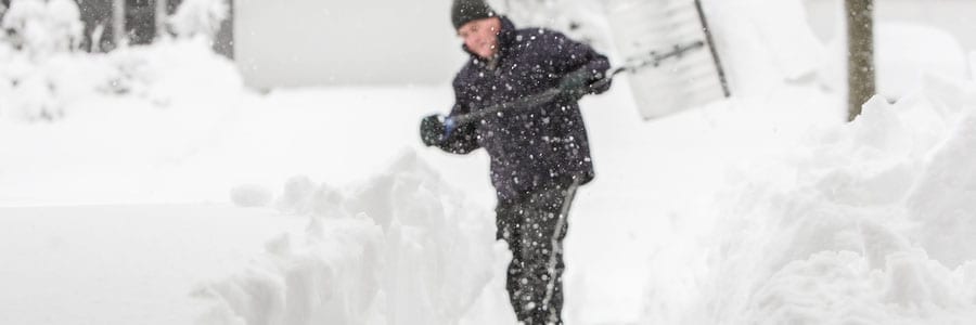 A person wearing winter clothing shovels snow in a snowy environment. Snowflakes are falling, and the scene is filled with deep snow.