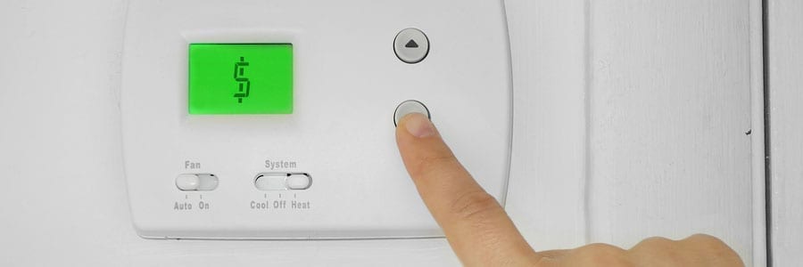 A finger presses a button on a wall-mounted thermostat with a green screen displaying a dollar sign.