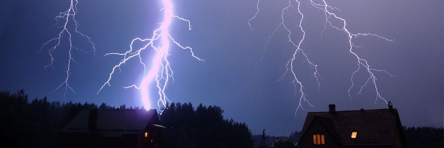 Lightning strikes near houses at night, illuminating the sky over a wooded area with bright bolts.