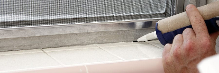 A person applies a line of caulk along the edge of a window frame and tiled surface using a caulking gun.