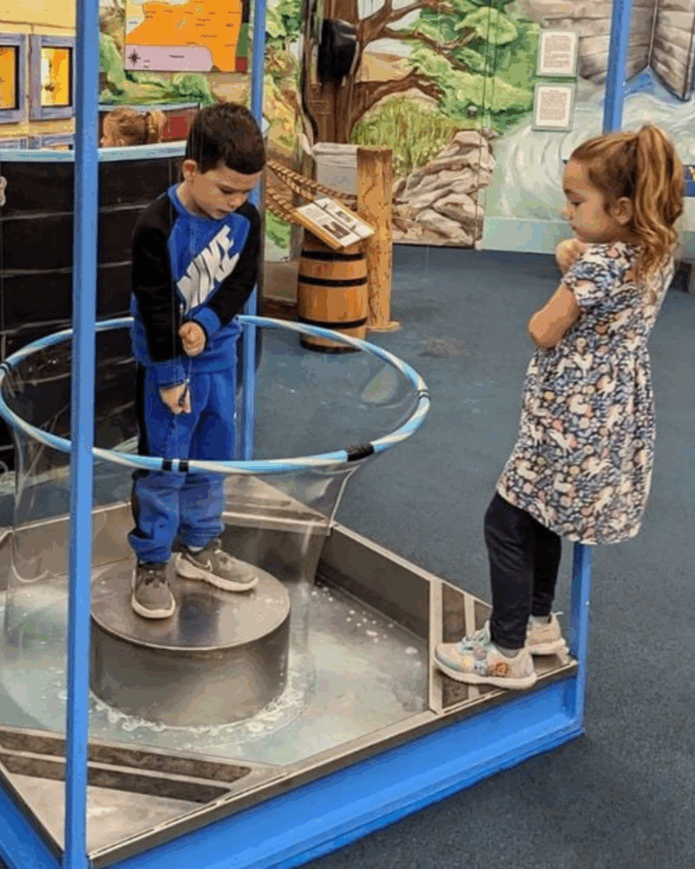 A boy stands inside a circular platform surrounded by a large bubble wand, while a girl watches nearby in a children's science exhibit sponsored by Auchinachie Cares.