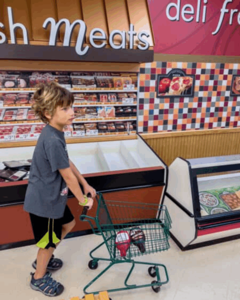 A young boy pushes a small shopping cart with groceries past the deli and meat section in a supermarket, showing that Auchinachie Cares about family moments.