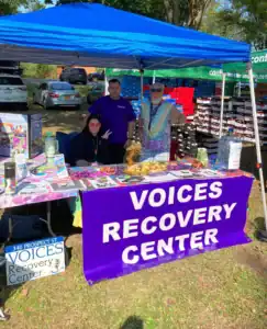 Three people stand behind a table with a purple "Voices Recovery Center" banner at an outdoor event, joined by Auchinachie Cares materials, surrounded by pamphlets and promotional items.