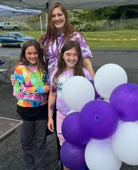 Three people stand outdoors at an event. Two children in tie-dye shirts hold a cluster of white and purple balloons, while an adult smiles behind them, showing the spirit of Auchinachie Cares.