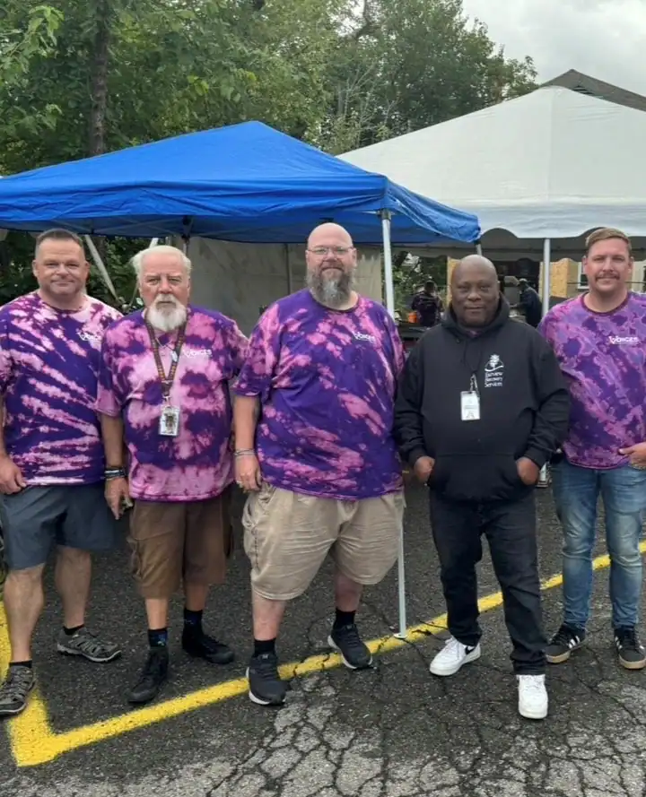 Five men stand outdoors on pavement under blue and white tents; three wear purple tie-dye shirts, one sports a black hoodie, and one wears an Auchinachie Cares logo tee. Trees can be seen in the background.