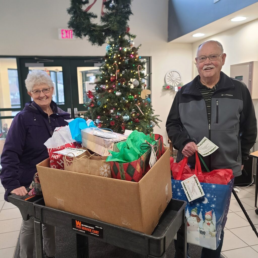 Two older adults stand indoors by a decorated Christmas tree, their cart full of gift bags and boxes. Both hold envelopes and smile warmly at the camera, embodying the spirit of giving with Auchinachie Cares.