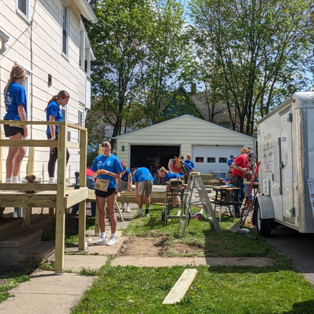 A group of people wearing blue shirts work on a house renovation project outdoors, assembling materials and tools near a garage and trailer on a sunny day as part of the Auchinachie Cares initiative.