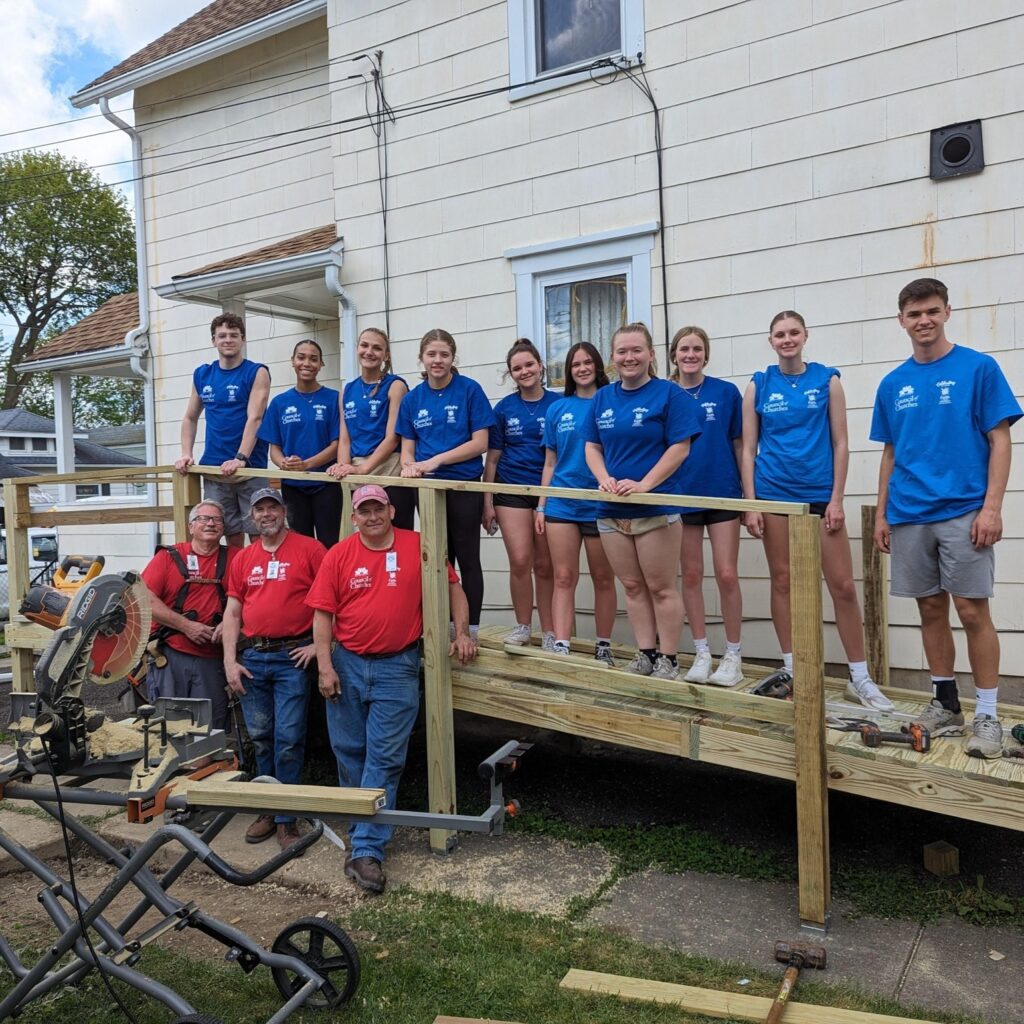 A group of people stands on a wooden ramp under construction, some in blue shirts and others in red, in front of a light-colored house. Tools and wood are scattered around as the Auchinachie Cares team works together.