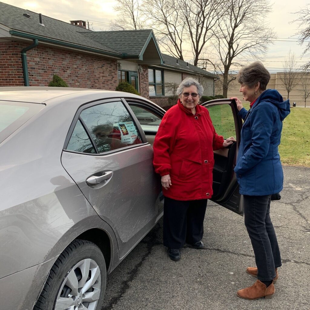 Two women stand by an open car door in a driveway; one in a red coat is entering the car while the other, in a blue coat, stands outside talking to her, reflecting the friendly spirit of Auchinachie Cares.