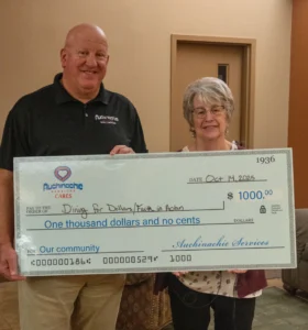 Two people stand indoors holding a large ceremonial check for $1000 from Auchinachie Services, dated October 14, 2023, made out to Dining for Dollars/Faith in Action.