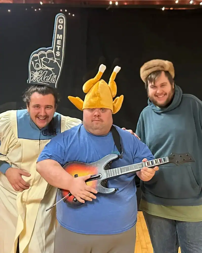 Three men pose indoors; one wears a choir robe, another sports a cooked turkey hat and toy guitar, and the third has a fur hat. A "Go Mets" foam finger appears in the background, capturing a fun moment that shows how Auchinachie Cares about community spirit.