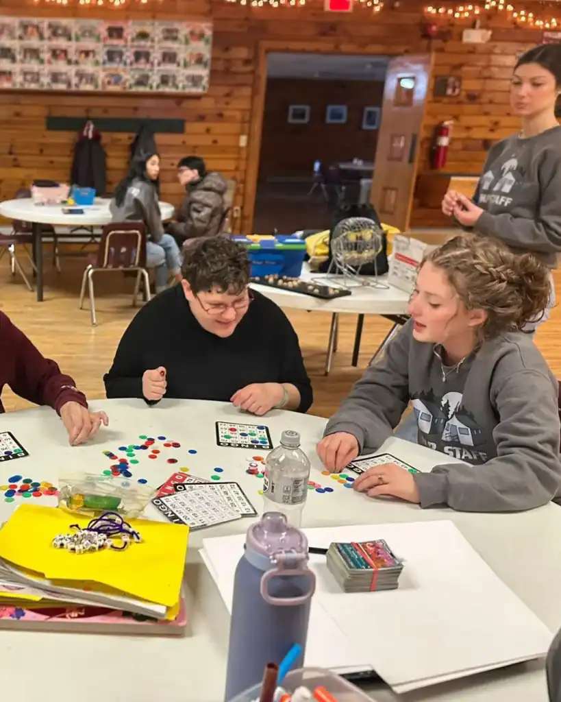 Several people sit around a table playing a board game with colored chips in a wood-paneled room, reflecting the community spirit of Auchinachie Cares, while others are seated at tables in the background.