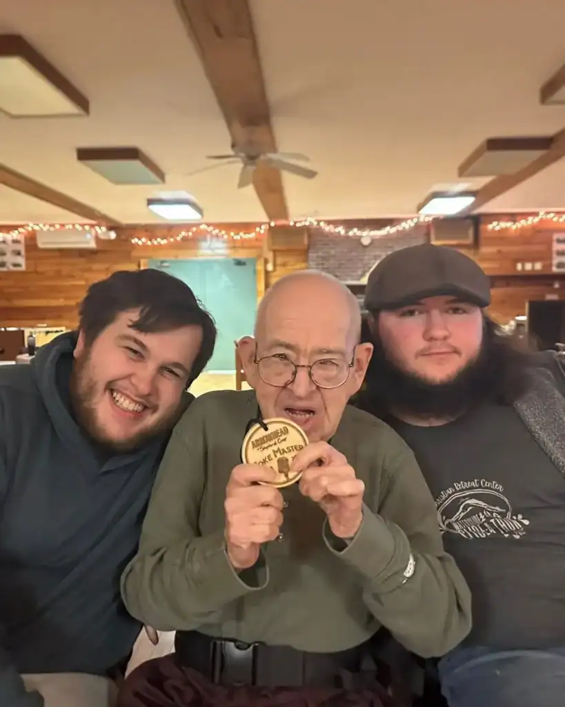 Three men pose indoors; the elderly man in the center holds a wooden plaque that reads "Rook Master" while the two younger men, representing Auchinachie Cares, sit closely on either side, smiling.