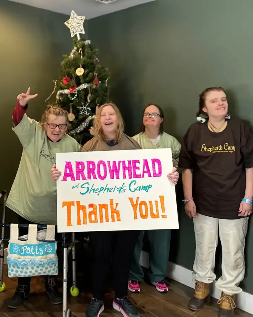 Four women stand in front of a decorated Christmas tree, smiling and holding a sign that reads "Arrowhead and Shepherds Camp Thank You!" as part of the Auchinachie Cares initiative.