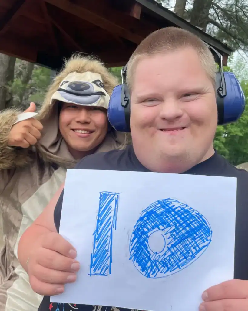 Two people outdoors; one in earmuffs smiles and holds a paper with "10," while someone in a sloth costume gives a thumbs up, showing their Auchinachie Cares spirit in the background.