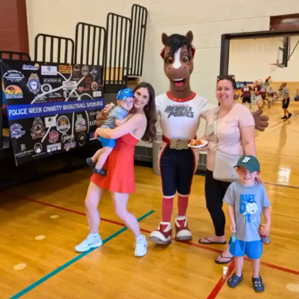 Two women and two children pose with a horse mascot in a gymnasium during a basketball event; an Auchinachie Cares sponsor board is displayed in the background.