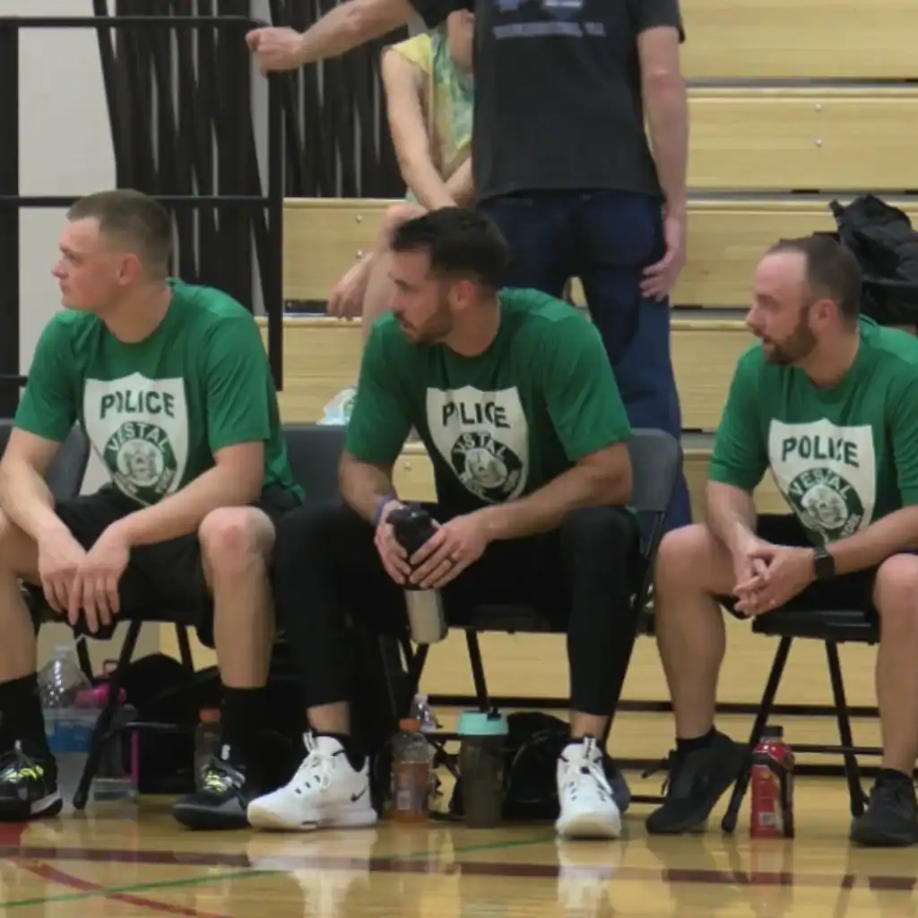 Three men in green "POLICE" t-shirts sit on chairs in a gymnasium, with sports bottles at their feet. The scene, part of an Auchinachie Cares event, shows bleachers and other people visible in the background.