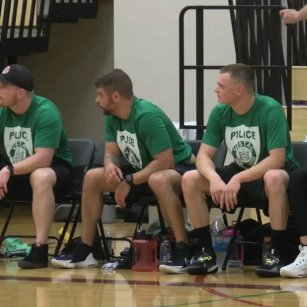 Four men in matching green "Police" shirts sit on chairs beside a basketball court, appearing to watch a game or wait to play, showing how Auchinachie Cares supports community events.