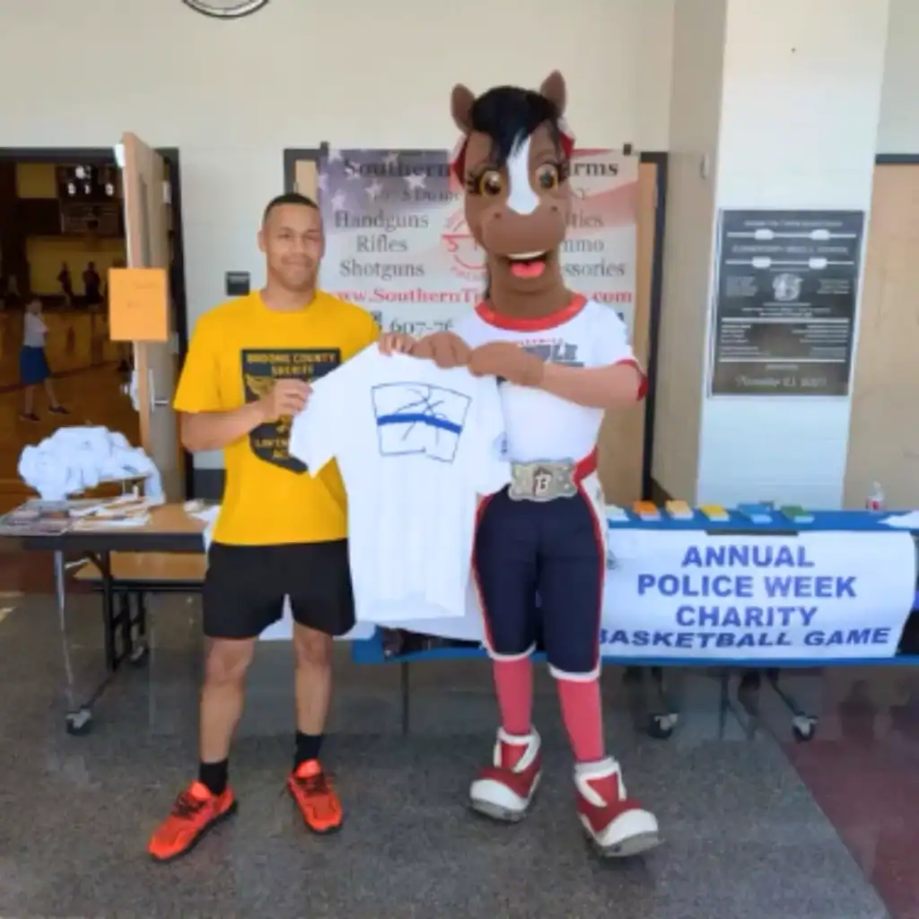 A man and a horse mascot hold up a white T-shirt in front of a table with a sign reading "Annual Police Week Charity Basketball Game," proudly showing support for Auchinachie Cares.