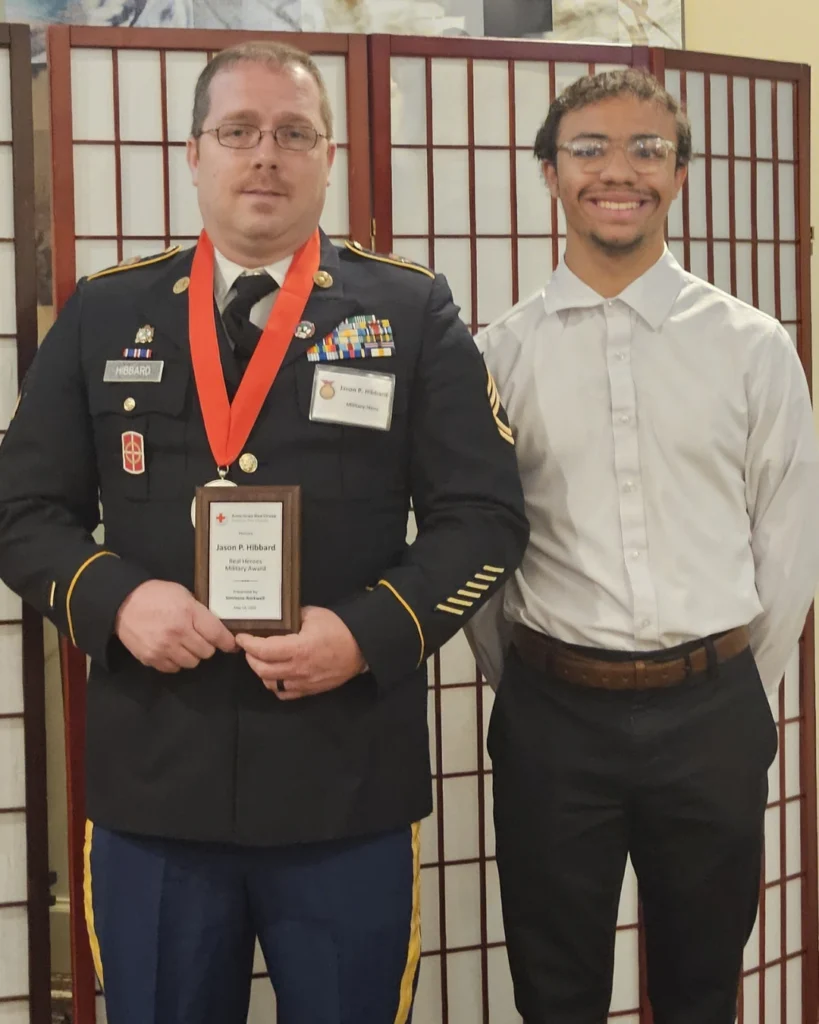 Two men stand side by side; one in a military uniform holding a plaque and wearing a medal, the other in a white shirt and dark pants. Both face the camera against a folding screen, honoring achievement as part of the Auchinachie Cares initiative.