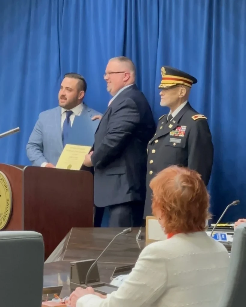 Three men stand together, one in a military uniform with medals, another holding a certificate printed with "Auchinachie Cares," posing for a photo at a podium in front of a blue curtain during an official event.