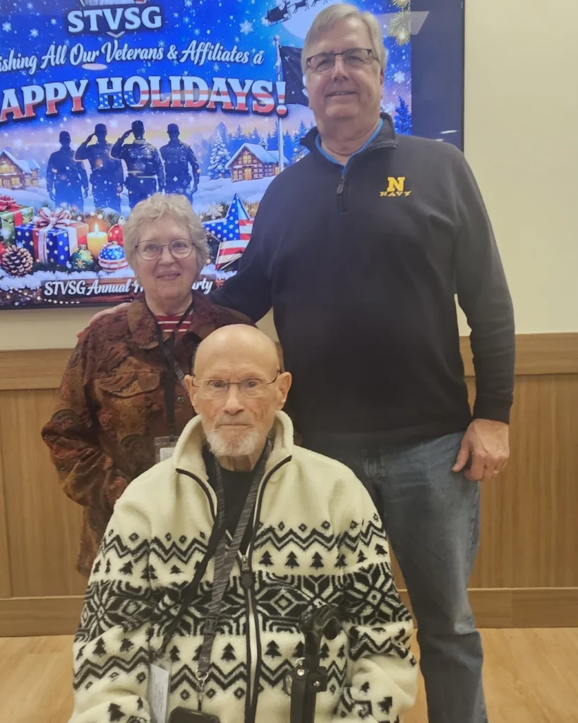 Three older adults pose indoors in front of a holiday-themed display that reads "Happy Holidays" with festive graphics, a message for veterans, and the supportive spirit of Auchinachie Cares.
