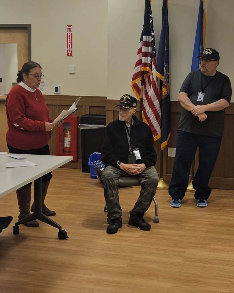 A woman reads from a paper to two men, one seated and one standing, in a room with flags, a fire extinguisher, and an Auchinachie Cares banner in the background.