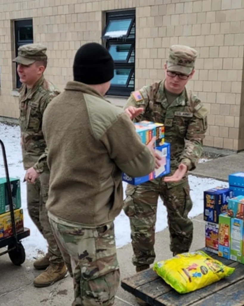 Three people in military uniforms move and stack boxes of food and supplies outdoors near a building on a snowy day, demonstrating the Auchinachie Cares spirit.
