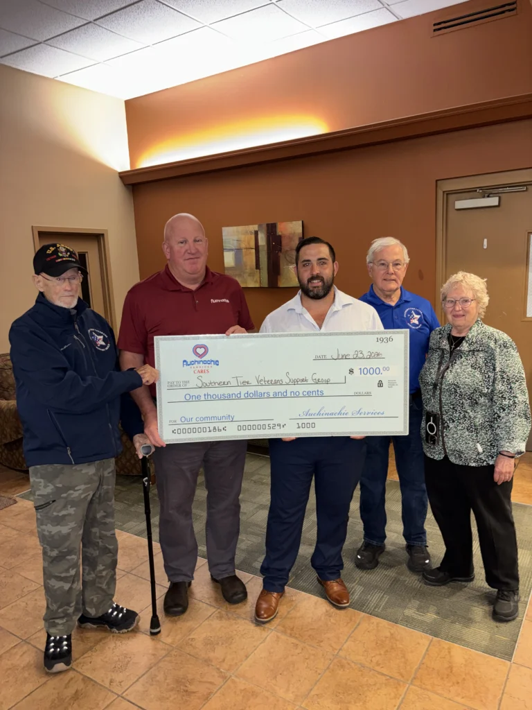 Five adults stand indoors holding a large ceremonial check for $1,000 made out to the Veterans Support Group, presented by Auchinachie Cares.