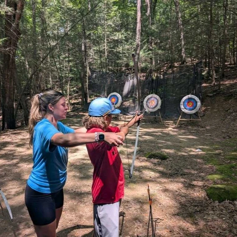 An adult instructs a child on archery in a wooded outdoor setting, with three target boards in the background—showcasing how Auchinachie Cares about fostering skills and confidence in future generations.