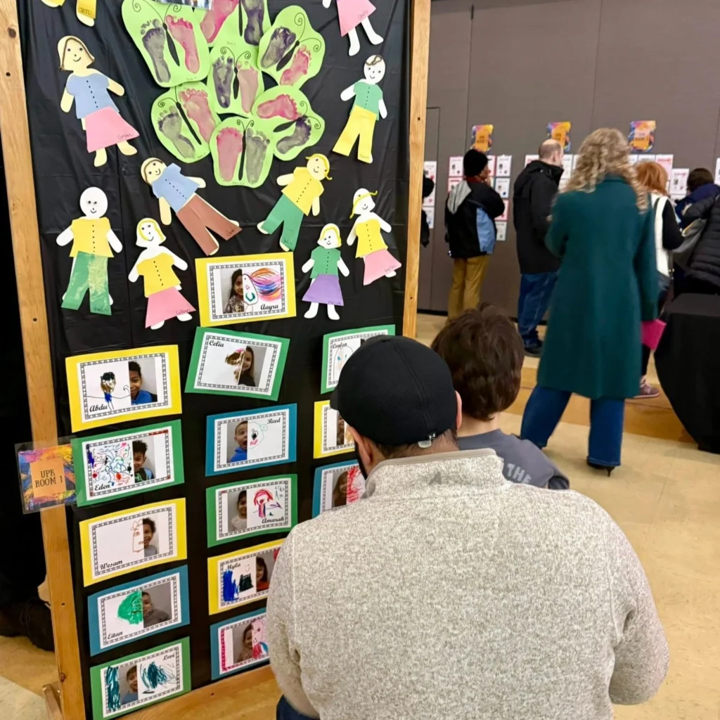 Adults and children view a display of children's photos, drawings, and paper cutouts at an indoor event supported by Auchinachie Cares. Other people are visible in the background.