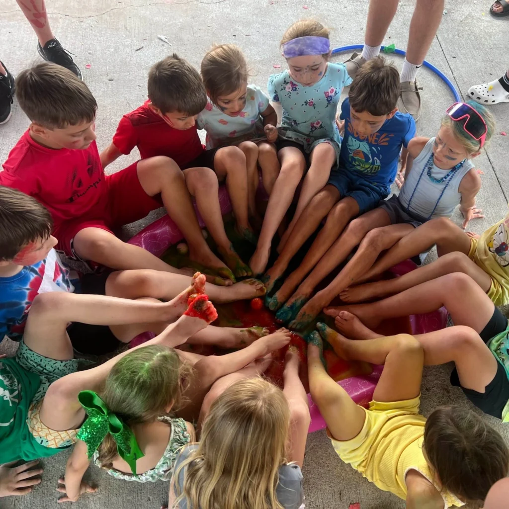 A group of children sitting in a circle with their feet in a small plastic pool, engaging in a messy activity involving colorful substances—an afternoon of fun made possible by Auchinachie Cares.
