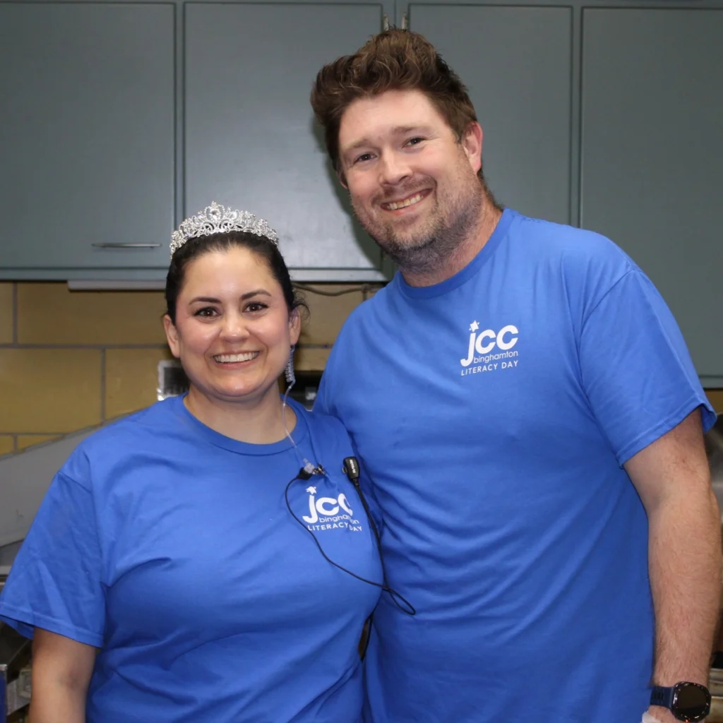 Two people wearing matching blue "JCC Literacy Day" shirts smile at the camera; one is wearing a tiara. They stand in a room with grey cabinets, celebrating community spirit supported by Auchinachie Cares.